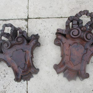 May include: Two ornate, dark brown wooden wall brackets with intricate carvings. Each bracket features a central shield-shaped design and a swirling, scroll-like top.