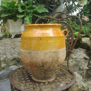 May include: A large, yellow and brown glazed ceramic pot with two handles. The pot is sitting on a metal table with a woven pattern.