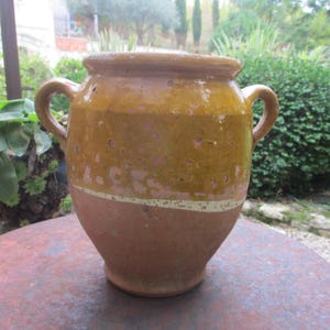 Könnte beinhalten: Ein großer, brauner Tonkrug mit zwei Henkeln. Der Krug hat einen weißen Streifen um die Mitte und ist in einem gefleckten Braun und Gelb glasiert.