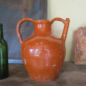 May include: An orange ceramic jug with two handles and a green glass wine bottle are displayed on a wooden surface. A small, reddish-brown relief sculpture is also visible. The background features a blue and brown wall.