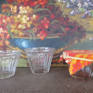 May include: Three clear glass jars with fluted sides. The jar on the right is covered with a colorful fabric lid with a rubber band.