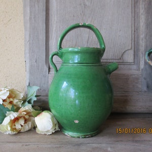 Op de afbeelding: Een groene geglazuurde keramische kan met een handvat. De kan heeft een rond lichaam en een tuit. De kan staat op een houten oppervlak.