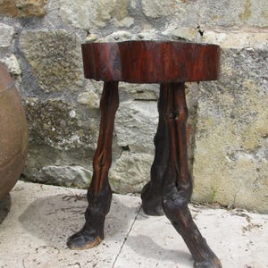 Puede incluir: Un taburete de madera artesanal con una parte superior circular de color marrón oscuro y tres patas talladas intrincadamente. Las patas tienen una forma natural y orgánica, lo que le da al taburete una apariencia artística única. El taburete está hecho de madera marrón oscuro.