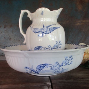 Puede incluir: Un juego de jarra y lavabo de porcelana blanca con diseños florales y de pájaros azules. La jarra tiene un asa y un pico. El lavabo es redondo y tiene un borde festoneado.