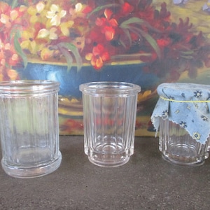 Może przedstawiać: Trzy przezroczyste szklane słoiki z żebrowanymi bokami. Słoiki są różnych rozmiarów, a najmniejszy słoik ma niebiesko-białą pokrywkę z tkaniny.