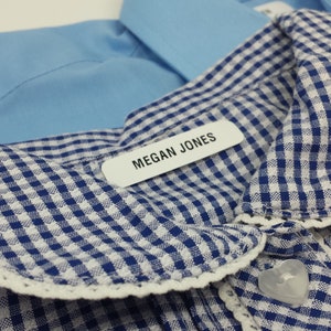 Op de afbeelding: Close-up van een blauw-witte geruite kraag met witte kanten rand. Een wit label met de naam "MEGAN JONES" is aan de kraag bevestigd. Een lichtblauw overhemd is zichtbaar op de achtergrond.