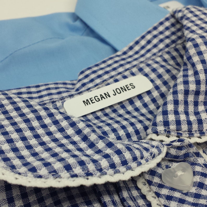 Op de afbeelding: Close-up van een blauw-witte geruite kraag met witte kanten rand. Een wit label met de naam "MEGAN JONES" is aan de kraag bevestigd. Een lichtblauw overhemd is zichtbaar op de achtergrond.