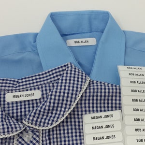 Op de afbeelding: Afbeelding van kledinglabels met de namen "BOB ALLEN" en "MEGAN JONES" erop gedrukt. De labels zitten op een lichtblauw overhemd en een blauw-wit geruit overhemd. Een stapel extra labels is zichtbaar.