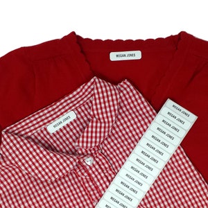 Op de afbeelding: Rood vest en rood-wit geruit overhemd met naamlabels "MEGAN JONES". Een vel met bijpassende labels is ook zichtbaar. Ideaal voor het labelen van kinderkleding.