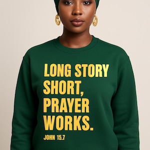 May include: A dark green sweatshirt with the text "LONG STORY SHORT, PRAYER WORKS." in yellow block letters. The text "JOHN 15.7" is below. The person in the image is wearing a matching green head covering and gold hoop earrings.