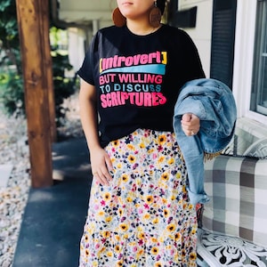 May include: A woman wearing a black t-shirt with the text "[Introvert] But Willing To Discuss Scriptures" in colorful letters. She is also wearing a floral maxi skirt and a denim jacket.