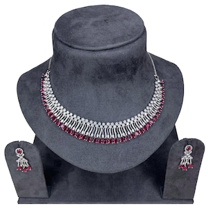 May include: A silver necklace and earring set displayed on a dark grey bust. The necklace has a curved design with alternating clear and ruby-coloured stones. The earrings mirror the necklace's design, with a ruby-coloured stone at the centre and dangling accents.
