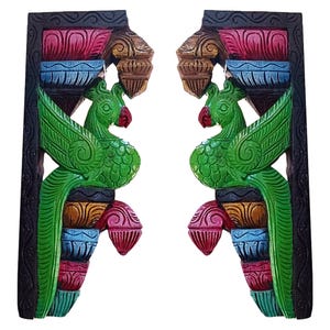 May include: Two matching, hand-carved wooden wall brackets. Each bracket features a green parrot with red accents, set against a backdrop of colorful, stacked architectural elements in shades of pink, blue, and gold. The brackets have a black border.