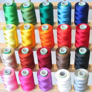Puede incluir: Una colección de 24 carretes de hilo en varios colores, incluyendo verde, azul, amarillo, rojo, naranja, marrón, negro, rosa, morado, blanco y gris. Los carretes están dispuestos en filas en un estante de madera.
