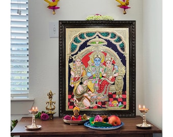 Pode incluir: Uma pintura hindu colorida e emoldurada retratando divindades em um estilo tradicional. A obra de arte apresenta figuras com tons de pele azuis e amarelos, adornadas com joias e roupas ornamentadas. A pintura é ambientada em um fundo escuro com detalhes dourados. A cena está em uma mesa de madeira com velas e itens religiosos.
