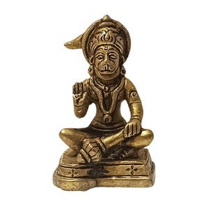 Op de afbeelding: Een messing beeld van een zittende godheid met een aapachtig gezicht, met een kroon en sierlijke sieraden. De figuur bevindt zich in een meditatieve pose, met één hand geheven in een zegenend gebaar. Het beeld staat op een vierkante basis met decoratieve patronen.