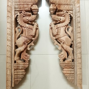 Peut inclure: Deux supports en bois sculptés de manière complexe avec des motifs ornés représentant des créatures mythologiques. Les supports sont en bois brun clair et ont un style indien traditionnel.