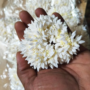 Puede incluir: Primer plano de un arreglo floral blanco sostenido en una mano. El arreglo es un grupo denso de pequeñas formas blancas en forma de pétalos con un centro amarillo. El fondo es blanco suave y desenfocado.