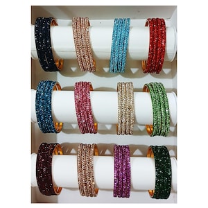 Puede incluir: Un conjunto de 10 pulseras coloridas con pedrería. Las pulseras están dispuestas en dos filas de cinco, con cada pulsera descansando sobre un soporte blanco. Las pulseras son de los siguientes colores: negro, dorado, azul, rojo, azul verdoso, rosa, dorado, verde, granate y verde oscuro.