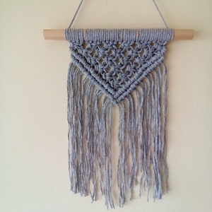 May include: A grey macrame wall hanging with a triangular shape and fringe. The hanging is attached to a wooden dowel rod.