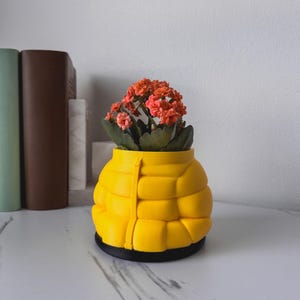 May include: A yellow planter shaped like a puffer jacket with a small plant with orange flowers growing inside.