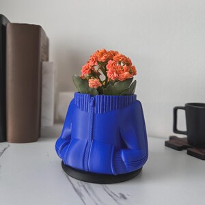 May include: A blue ceramic planter shaped like a zipped jacket with a small orange flower plant inside.