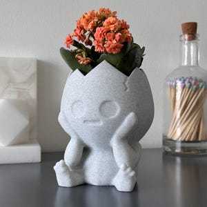 May include: A white ceramic planter shaped like a small person with a cracked egg on its head. The planter has a surprised expression and is holding its head in its hands. The planter is filled with a small orange flowering plant.
