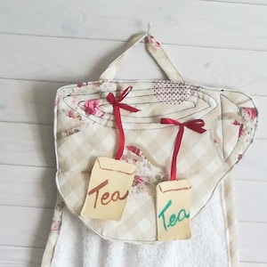 Può includere: Set di tre asciugamani da cucina a tema tè. Gli asciugamani sono realizzati in tessuto beige a quadri con dettagli floreali. Gli asciugamani presentano un design a tazza da tè con la parola "Tè" scritta su ogni asciugamano.