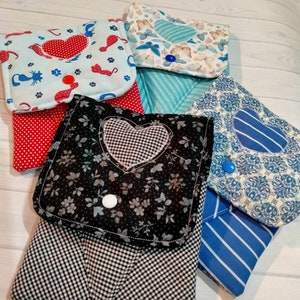 Può includere: Una collezione di pochette in tessuto fatte a mano con vari motivi. I disegni includono cuori, fiori, farfalle e strisce. Ogni pochette ha una chiusura con bottone ed è realizzata in tessuto di cotone colorato. Le pochette sono disposte su una superficie di legno chiaro.