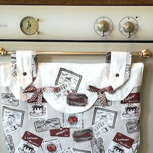 Può includere: Un organizzatore da cucina in tessuto bianco con un'asta in metallo marrone e un nastro a pois marroni. L'organizzatore ha un tema caffè vintage con etichette marroni e rosse che dicono "Vintage Coffee", "Vintage Breakfast", "Pure Cider" e "Coffee".