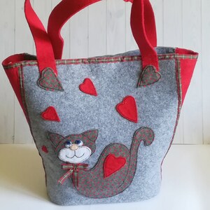 Può includere: Una borsa tote in feltro grigio con manici rossi e un design di gatto rosso. Il gatto ha un viso bianco e occhi blu. La borsa è decorata con cuori rossi.