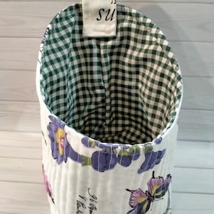 Può includere: Un organizzatore sospeso in tessuto bianco con una fodera a quadri verde e bianca. L'organizzatore ha una stampa floreale con farfalle e il testo "56'6" stampato sul davanti. L'organizzatore ha un anello in alto per appenderlo.