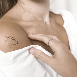 May include: A single-line drawing of a dog's head is tattooed on a person's shoulder.