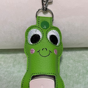 May include: A green frog-shaped hand sanitiser holder with a silver clip. The holder has embroidered details, including large eyes, pink cheeks, and a smiling mouth. A white hand sanitiser bottle is visible.