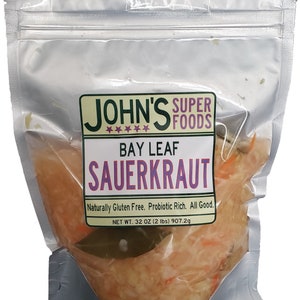 May include: A clear plastic bag filled with sauerkraut. The bag has a label that reads "John's Super Foods Bay Leaf Sauerkraut" and "Naturally Gluten Free. Probiotic Rich. All Good." The label also states "Net Wt. 32 oz (2 lbs) 907.2g".