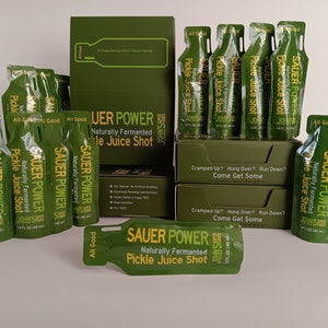 May include: A box of green and yellow Sauer Power Naturally Fermented Pickle Juice Shots. The box is surrounded by individual pouches of the pickle juice shot. The pouches are green and yellow and have the text "Sauer Power" and "Naturally Fermented Pickle Juice Shot" on them. The box has the text "Cramped Up? Hung Over? Run Down? Come Get Some" on it.