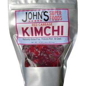 May include: A silver pouch with a clear window showing red cabbage kimchi. The label reads "John's Super Foods Red Cabbage Kimchi" and "Naturally Gluten Free. Probiotic Rich. All Good." The net weight is 16 oz (1 lbs) 453.6g.