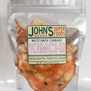 May include: A silver resealable pouch filled with white napa cabbage kimchi. The label reads "John's Super Foods White Napa Cabbage Kimchi" and "Naturally Gluten Free. Probiotic Rich. All Good." The net weight is 32 oz (2 lbs) 907.2g.