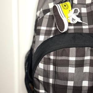 Puede incluir: Una mochila a cuadros en blanco y negro con un peque&ntilde;o llavero de zapatilla amarillo y blanco adjunto. El llavero tiene un logotipo de estrella y las palabras "The original" impresas. La mochila tiene un ribete negro y un bolsillo lateral de malla.