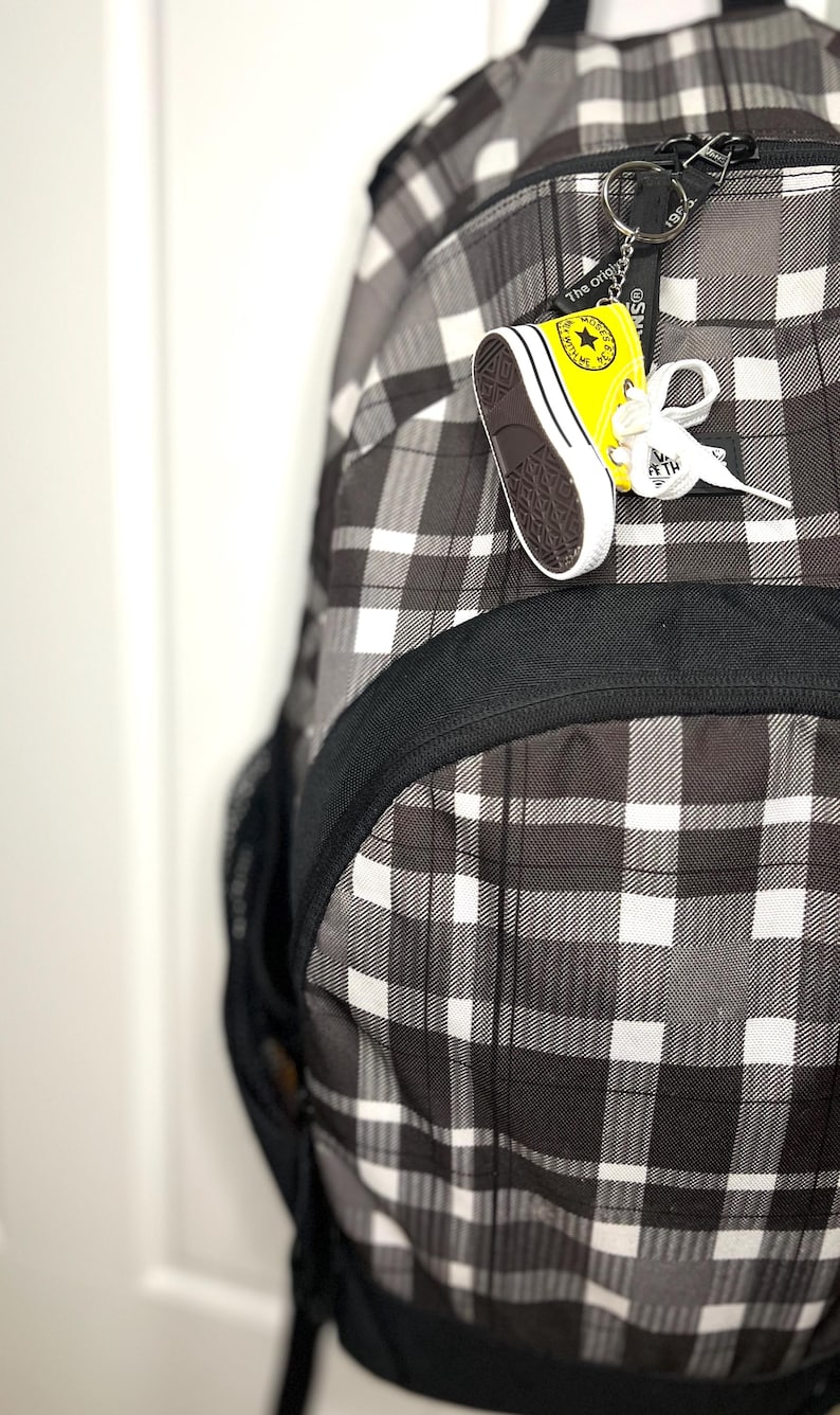 Puede incluir: Una mochila a cuadros en blanco y negro con un peque&ntilde;o llavero de zapatilla amarillo y blanco adjunto. El llavero tiene un logotipo de estrella y las palabras "The original" impresas. La mochila tiene un ribete negro y un bolsillo lateral de malla.