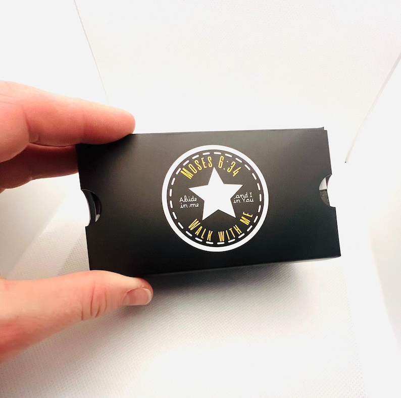 Puede incluir: Una caja de regalo negra con un dise&ntilde;o circular blanco y dorado con una estrella y el texto "MOSES 6:34, Abide in me, and I in You, WALK WITH ME". La caja est&aacute; sostenida por una mano.