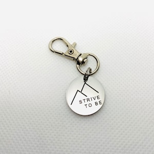 May include: Silver keychain with a round tag that says "STRIVE TO BE" with a mountain graphic.