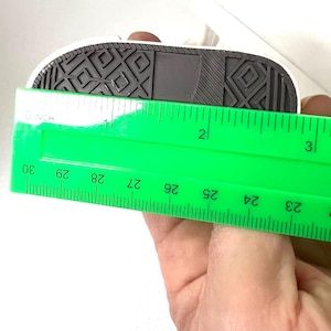 Puede incluir: Una suela de zapatilla blanca y negra en miniatura se mide con una regla verde. La suela mide aproximadamente 7,6 cm de largo. La suela tiene un patr&oacute;n texturizado negro y una parte superior blanca.
