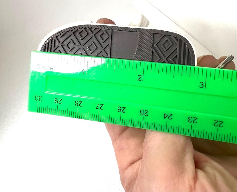 Puede incluir: Una suela de zapatilla blanca y negra en miniatura se mide con una regla verde. La suela mide aproximadamente 7,6 cm de largo. La suela tiene un patr&oacute;n texturizado negro y una parte superior blanca.