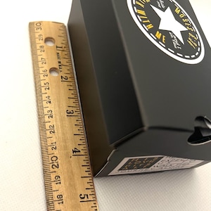 Puede incluir: Caja negra con un logotipo de estrella blanca y el texto "Walk with Me". La caja mide aproximadamente 10 cm de alto. Una regla de madera est&aacute; al lado de la caja.