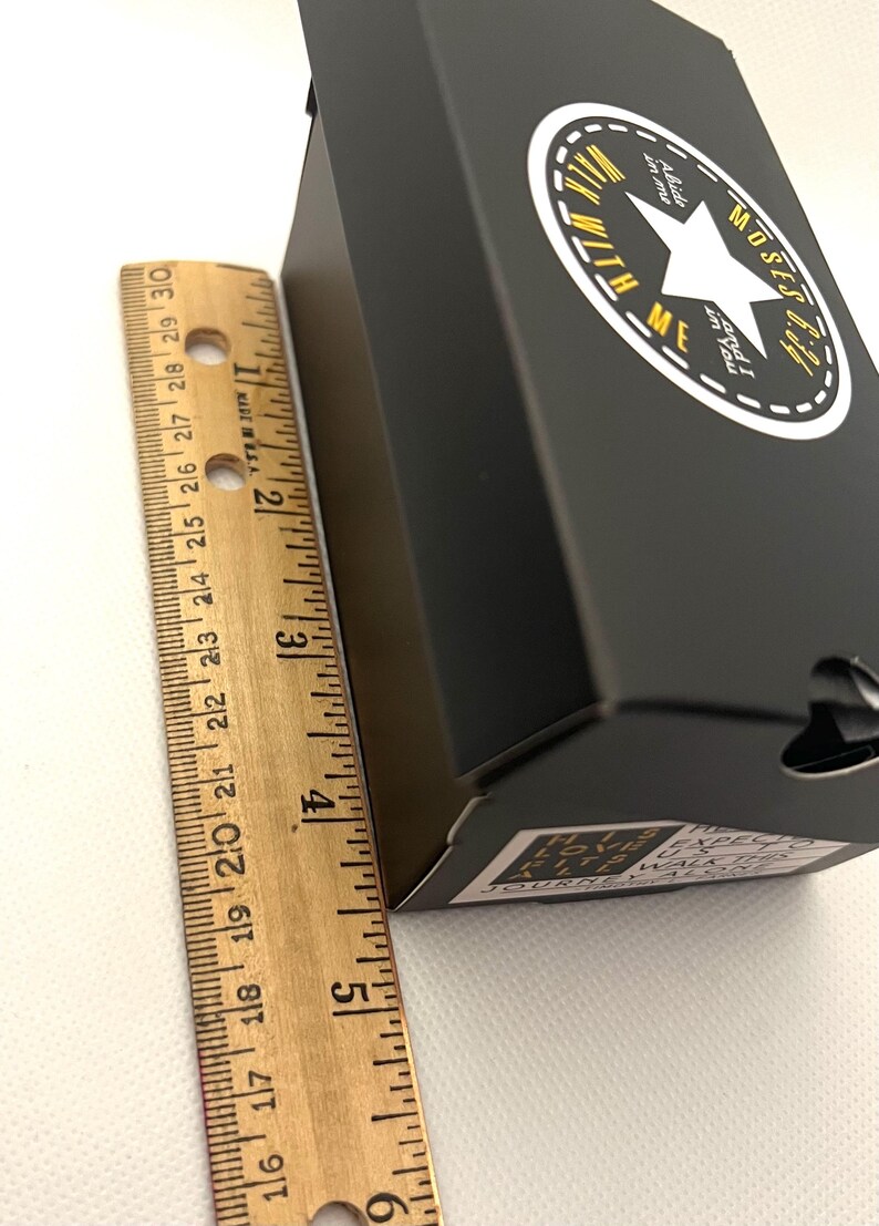Puede incluir: Caja negra con un logotipo de estrella blanca y el texto "Walk with Me". La caja mide aproximadamente 10 cm de alto. Una regla de madera est&aacute; al lado de la caja.
