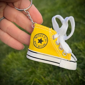 Puede incluir: Un llavero de zapatilla amarilla en miniatura con cordones blancos y un llavero plateado. La zapatilla tiene una suela negra y blanca y un emblema circular que dice "MOSES 6:34 WALK WITH ME".