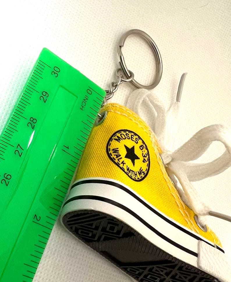 Puede incluir: Un llavero de zapatilla en miniatura amarilla con cordones blancos y suela negra. El llavero tiene un dise&ntilde;o en blanco y negro en el lateral con el texto "MOSES 6:34 WALK WITH ME". Una regla verde est&aacute; al lado del llavero.