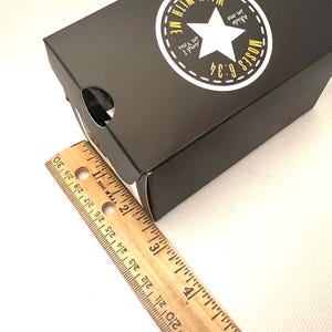 Puede incluir: Una caja negra con una estrella blanca y el texto "Abide in me and I in You, Moses 6:34." Una regla de madera est&aacute; al lado de la caja. La caja mide aproximadamente 25 cm de largo.