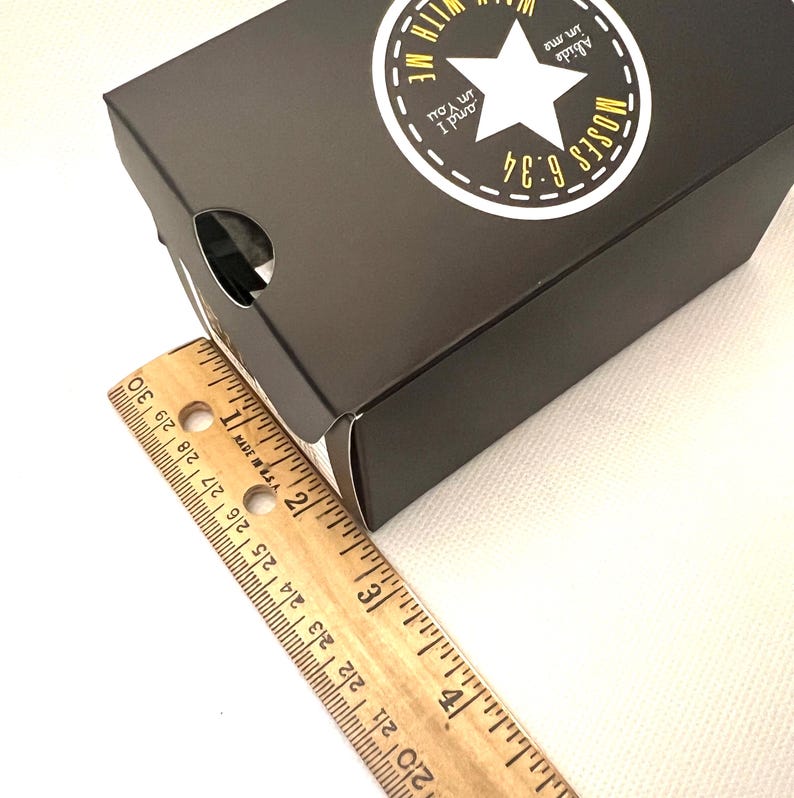 Puede incluir: Una caja negra con una estrella blanca y el texto "Abide in me and I in You, Moses 6:34." Una regla de madera est&aacute; al lado de la caja. La caja mide aproximadamente 25 cm de largo.